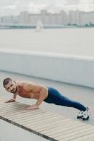 Flexible fit man does plank exercise on promenade has naked torso concentrated away prepares for workout practices yoga dressed in sportswear poses outdoor trains in morning. Sporty lifestyle photo