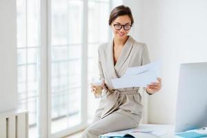 Pleased dark haired woman with smile, checks bills in documents, dressed beige business suit, drinks beverage, sits at desktop near big monitor, holds paper sheet with charts, poses near window photo