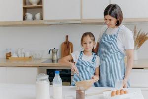 Smiling mother and daughter prepare tasty cookies, girl whisks ingredients in bowl with beater, helps mum at kitchen, prepare festive dinner, modern home interior. Family, cooking, lifestyle concept photo