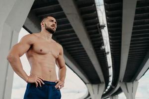 Strong man with naked torso keeps hands on waist, has muscular body after long training, focused into distance, stands under bridge, thinks about how to be healthy and fit. Sporty man outdoor photo