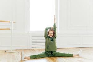 Full length shot of cheerful redhead woman raises hands, does splits, stretches muscles, sits on floor, stretches legs sideways, wears green tracksuit and sportshoes. Sport and active llifestyle photo