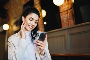 Smiling female with dark hair tied in pony tail wearing white clothes holding smartphone while listening to music on her electronic device. Stylish woman resting in cafe using app having smile on face photo