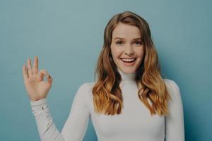 Beautiful excited joyful young lady with dyed light-brown hair gesturing ok symbol with fingers photo