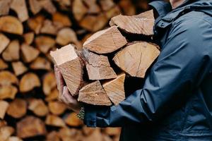 Close up of unrecognizable man dressed in jacket carries bundle of firewood, going to make fire outdoor. Winter is coming. photo