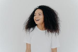 Portrait of beautiful curly adult woman turns gaze aside, has glad expression, smiles and laughs at something funny, dressed in white t shirt, models indoor, feels free, energized and relieved, photo