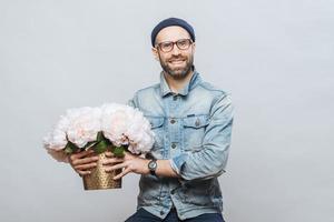 Happy delighted male buys bouquet of flowers for special occasion, wears denim jacket and denim shirt, isolated over white background. Joyful emotional man with present. Beautiful flowers for you photo