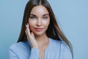 Gorgeous dark haired woman touches cheek gently, has direct gaze at camera, looks with tenderness, wears casual sweater, poses against blue background, has perfect glowing skin. Beauty, hair care photo