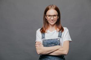 Photo of happy dark haired young woman keeps arms folded, smiles positively, wears eyewear, t shirt and denim overalls, isolated over grey background with free space for your information or text