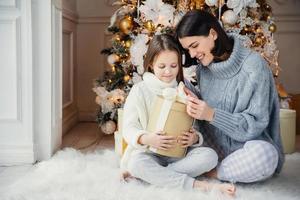 toma interior de una niña adorable y su madre sentadas con las piernas cruzadas, abren una caja de regalo envuelta, intrigan lo que hay allí, se sientan contra el año nuevo adornado o el árbol de navidad, tienen sonrisas sinceras y gentiles en las caras foto