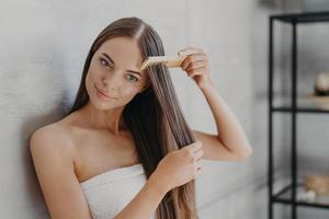 Young brunette woman brushes hair with comb after taking shower and applying hair care mask, wears minimal makeup, has healthy glowing skin after hygienic procedures, stands wrapped in towel photo