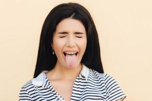 Portrait of beautiful woman makes grimace, keeps mouth wide opened and shows tongue, keeps eyes closed, wears casual clothes, poses against studio background. People and facial expressions concept. photo