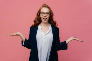 Half length shot of confused business lady raises palms, has hesitant facial expression, dressed in formal suit, spectacles, expresses bewilderment, isolated over pink background. People and doubt photo