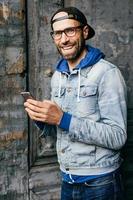 Stunning man with blue eyes, beard and broad smile wearing cap, denim shirt and eyeglasses holding smartphone isolated over old wall background. Pleased man in stylish clothes using cell phone photo