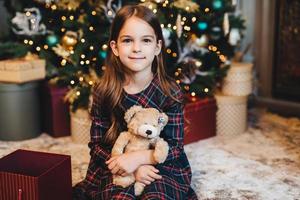 Small female child with appealing appearance, holds teddy bear, recieves present from parents, sits near Christmas tree. Adorable girl being glad to get gift on New Year. Christmas time. photo