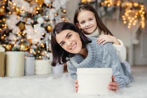 Children, family and celebration concept. Adorable female in knitted sweater holds white present box and small kid stands behind her back, have good mood before New Years Eve. Waiting miracle photo