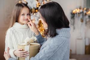 Portrait of adorable little kid and mother, touches her little nose, gives wrapped gift box, prepares little surpise for daughter, gives present near decorated New Year tree. Christmas and New Year