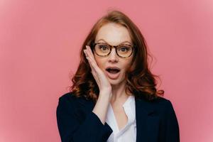 Really no way Shocked red haired woman touches cheek, opens mouth widely, looks surprisingly at camera, finds out shocking news, wears spectacles, elegant formal suit, isolated on pink background photo