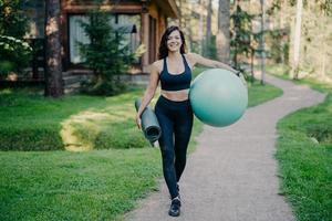 Outdoor shot of happy sporty brunette woman walks on path near forest, holds rolled up karemat and fitness ball, dressed in cropped top, leggings and sneakers, has regular trainings in open air photo