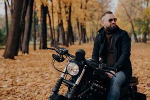 Outdoor shot of pensive male biker keeps gaze aside, has thoughtful look, wears sunglasses, sweater, black jacket and jeans, poses on motorbike outdoor in autumn park. Freedom, lifestyle, hobby photo