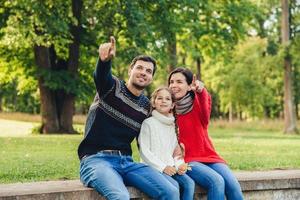 Father and mother sit between their daughter against trees or nature background, show something to her, indicate with fore fingers. Smiling parents show bird to little beautiful girl. Parenthood photo