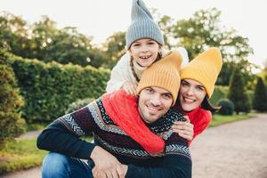 Outdoor portrait of handsome man give piggyback to his wife and daughter, wear warm clothes, have happy expressions, support each other. Family embrace outdoors, smile pleasantly into camera photo