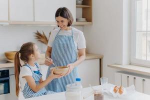 Indoor shot of caring mother with happy smile, cooks together with daughter in kitchen, prepare holiday pie, have pleasant talk, stand near table with different ingredients. Small helper helps mommy photo