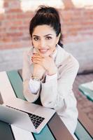 Luxurious brunette female with charming smile having pleasant look into camera while sitting at cafe in front of opened laptop chatting with friends or her boyfriend. People and technology concept photo