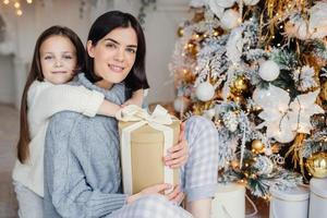 una pequeña niña agradecida abraza a su madre que le dio un regalo, pasan un tiempo maravilloso e inolvidable juntos, celebran la navidad. mujer morena e hija buscan regalos bajo el abeto foto