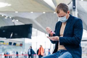 Photo of man in medical face mask, uses disinnfectant spray for hands, protects from coronavirus in crowded place, poses in airport, waits for flight. Health care concept. Sanitation, virus prevention
