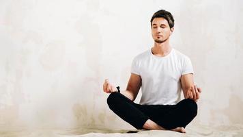 Young man sits in a meditative pose on sand against a wall background photo