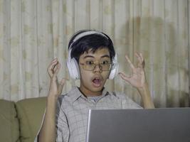 Teenage kid playing game console wearing headphone with raise hand after winning the game while sitting on sofa in living room. Online gaming concept. photo