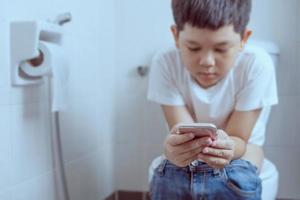 Asian boy sitting on toilet bowl while playing mobile phone - health problem concept photo