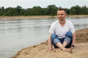 Image of a lonely positive and smiling man sitting on the river bank photo