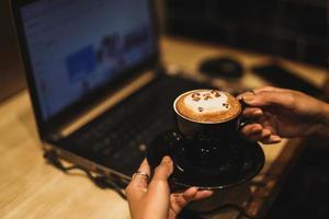 Female freelancer working online at computer while holding latte art coffee mug. Concept of networking and occupation. photo