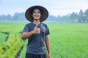 atractivo joven agricultor asiático alegre de pie y sonriendo mostrando el pulgar hacia arriba en el campo de arroz. concepto de agricultura moderna. foto