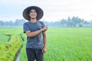 Attractive cheerful young Asian farmer standing and smiling pointing finger out to the rice field. Modern agriculture concept. photo