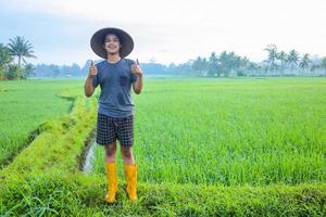 Attractive cheerful young Asian farmer standing and smiling showing thumbs up at the rice field. Modern agriculture concept. photo