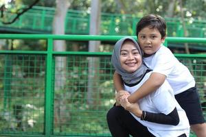 Portrait of Asian kid hugs mother from back, enjoying family time in the park. Bonding happy family concept. photo