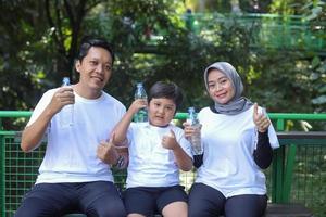 Happy Muslim family holding bottle with water while resting after sport workout outdoors in the public park. Parents and son smiling and giving thumbs up gesture. photo