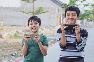 Two kids with gadgets. Brothers surfing the net or playing online games on smartphone and digital tablet outside while standing. Modern communication and gadget addiction concept. photo