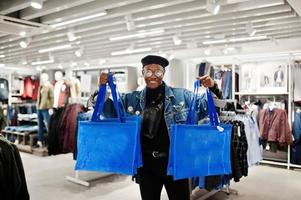 Stylish casual african american man at jeans jacket and black beret with fanny pack or waist bag holding blue shopping bags at clothes store. photo