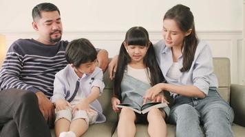 aziatische thaise familie, volwassen vader, moeder en kinderen geluk thuis wonen ontspannende activiteiten en samen een boek lezen, vrije tijd op de bank in een wit kamerhuis, heerlijk weekend, welzijn huiselijke levensstijl. video