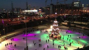 panela lenta de pessoas patinando no gelo em torno de uma árvore de natal em uma pista de patinação à noite com show de luzes video
