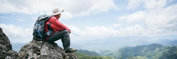 hombre joven haciendo senderismo en la roca superior, hombre de mochila mirando el hermoso valle de montaña a la luz del sol en verano, paisaje con hombre deportivo, colinas altas, bosque, cielo. viaje y Turismo. foto