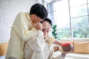 pareja gay asiática sorprende a su novio con un regalo y manos en los ojos en casa, concepto lgbtq. foto