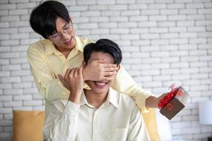 pareja gay asiática sorprende a su novio con un regalo y manos en los ojos en casa, concepto lgbtq. foto