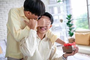 Asian gay couple surprise boyfriend with gift and hands on eyes at home, LGBTQ concept. photo