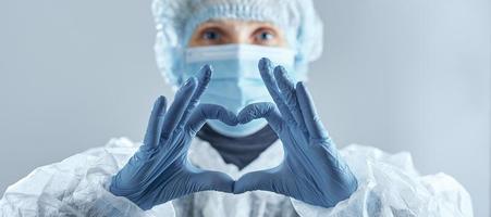 el trabajador médico borroso con máscara médica, guantes de goma y traje hace una señal de corazón. confrontando el coronavirus. foto