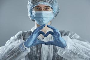 mujer joven con gafas con máscara médica y manos con guante de látex muestra el símbolo del corazón. foto