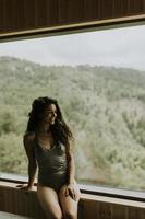 mujer joven en el centro de spa de bienestar sentada junto a la enorme ventana ancha disfrutando de la vista del bosque verde foto